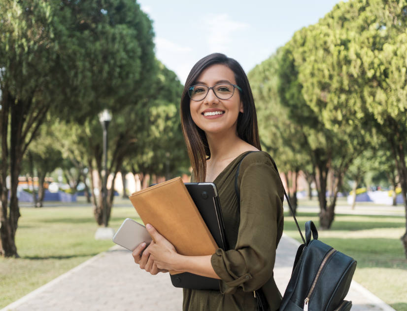 A female student smiles on campus, holding her books and phone and a bag